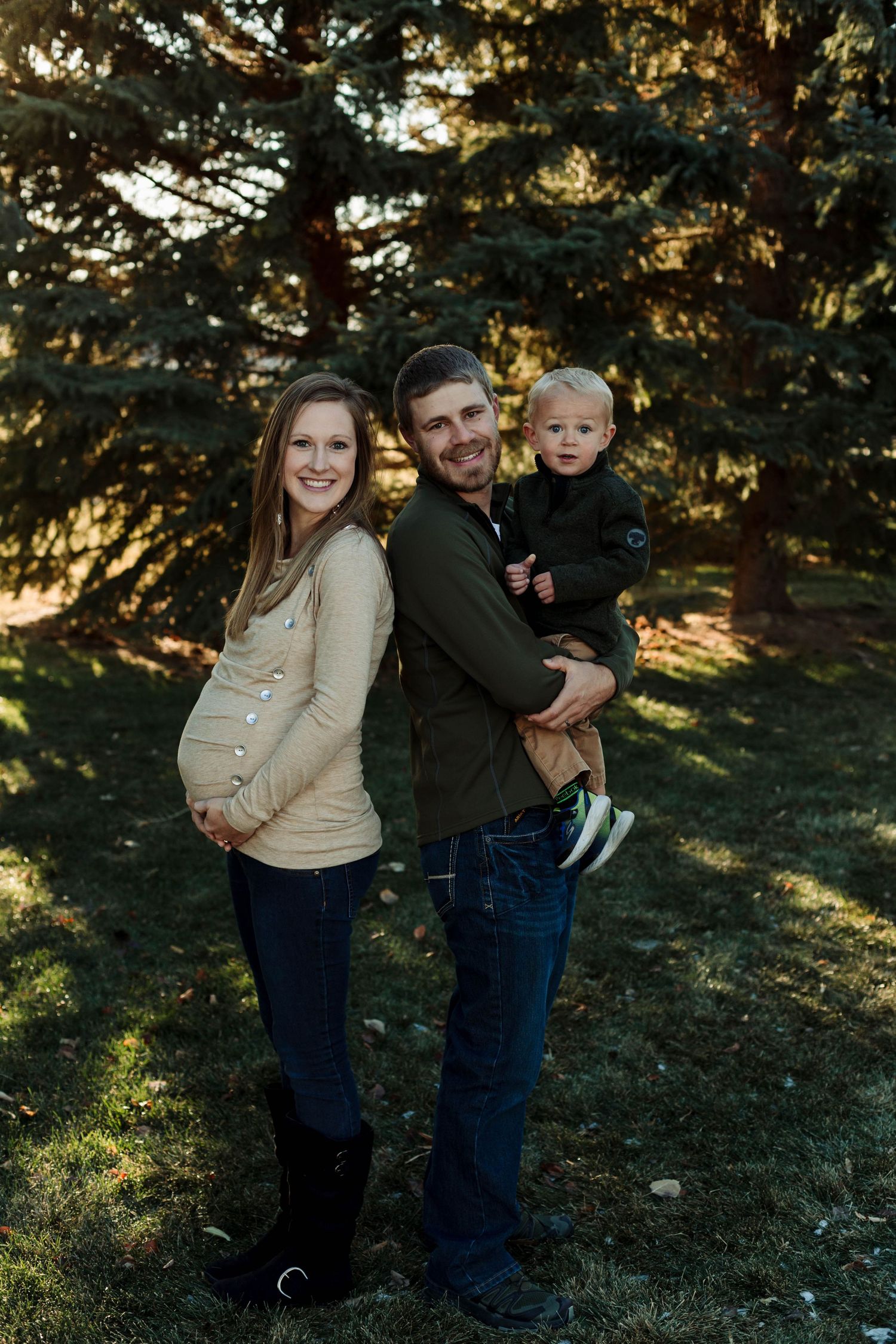 Family maternity photo in front of evergreen trees at golden hour.