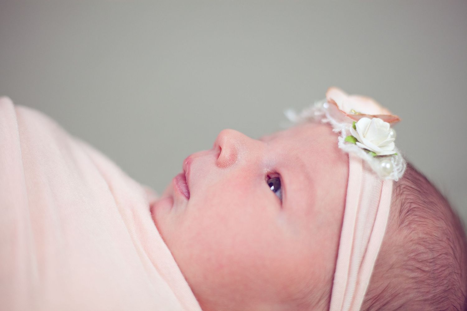 A newborn wearing a delicate white headband looking up against a soft pink background.