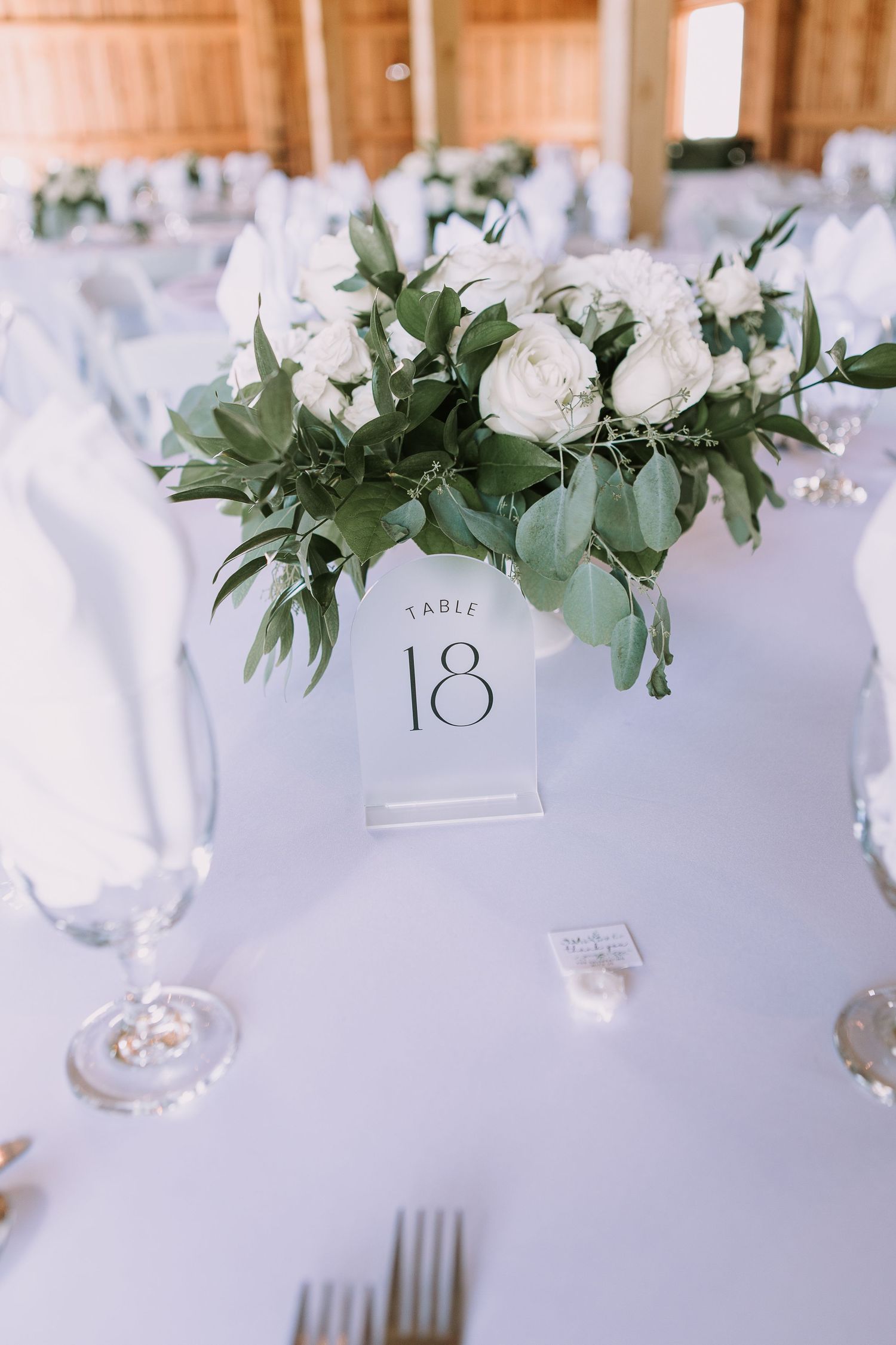 White rose and greenery centerpiece with table number 18 in an elegant barn reception setting.