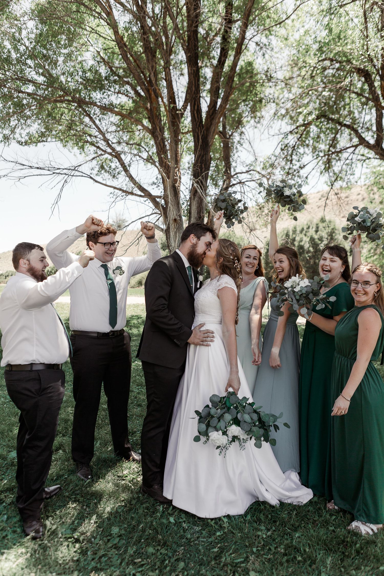 Joyful wedding party celebration moment under trees with bridesmaids in green dresses.