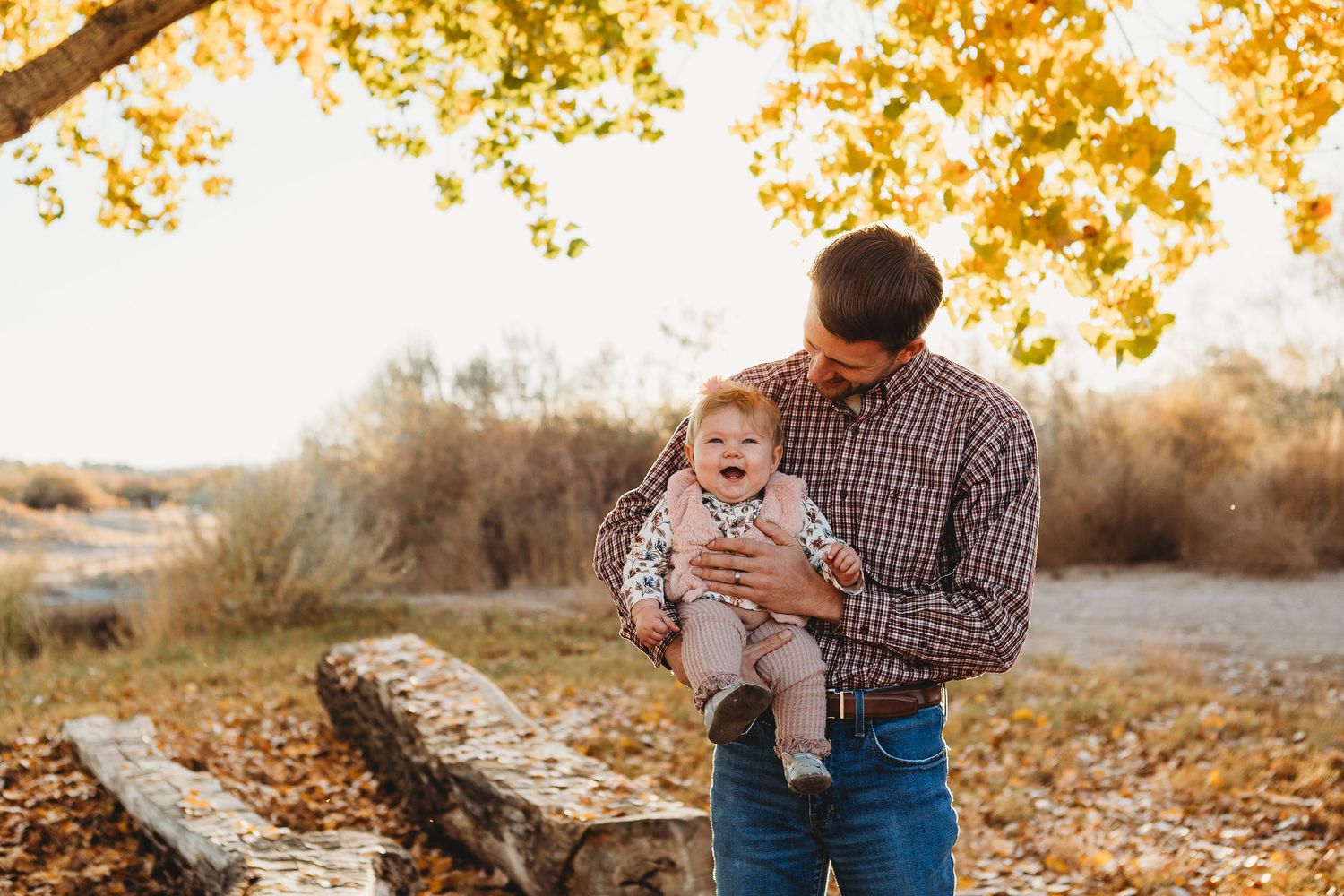 A plaid-shirted figure holds a small baby against a backdrop of autumn foliage and warm sunlight.