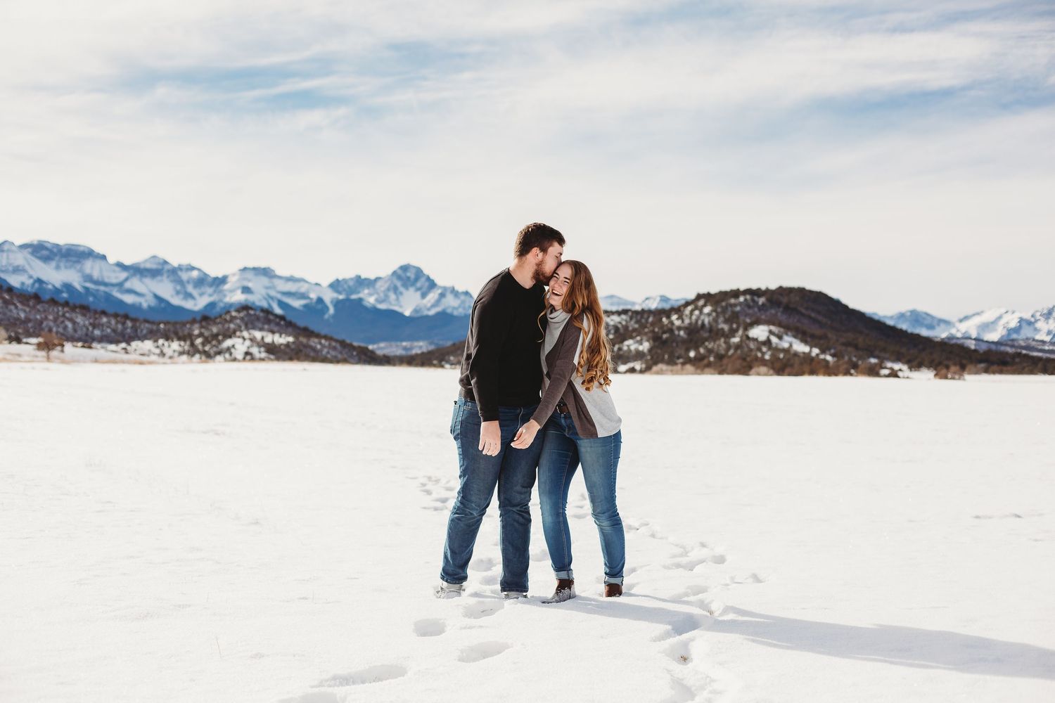 A romantic winter scene with a couple standing together in the snow with mountain views in the background.