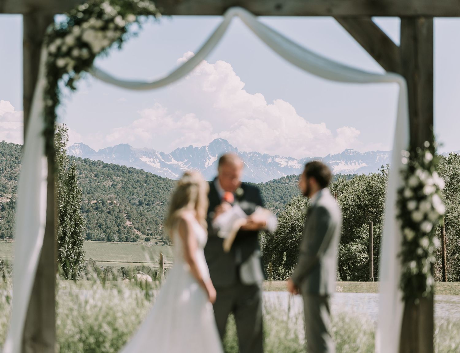 Wedding ceremony under wooden arch with mountain vista and white drapery.
