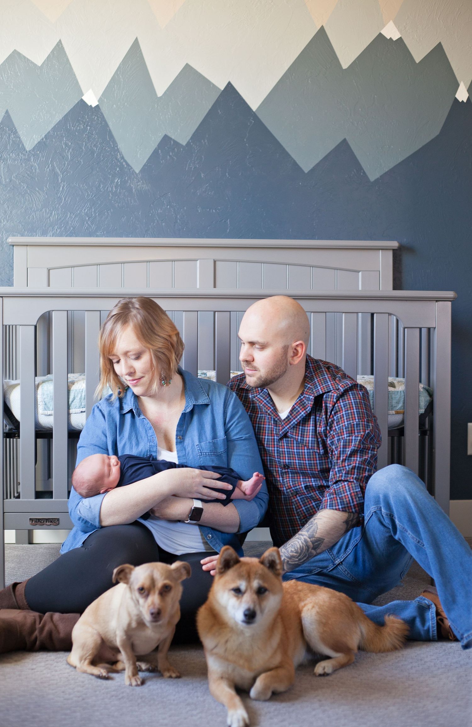 A nursery scene with mountain wall mural, grey crib, and two dogs resting by a couple with baby.