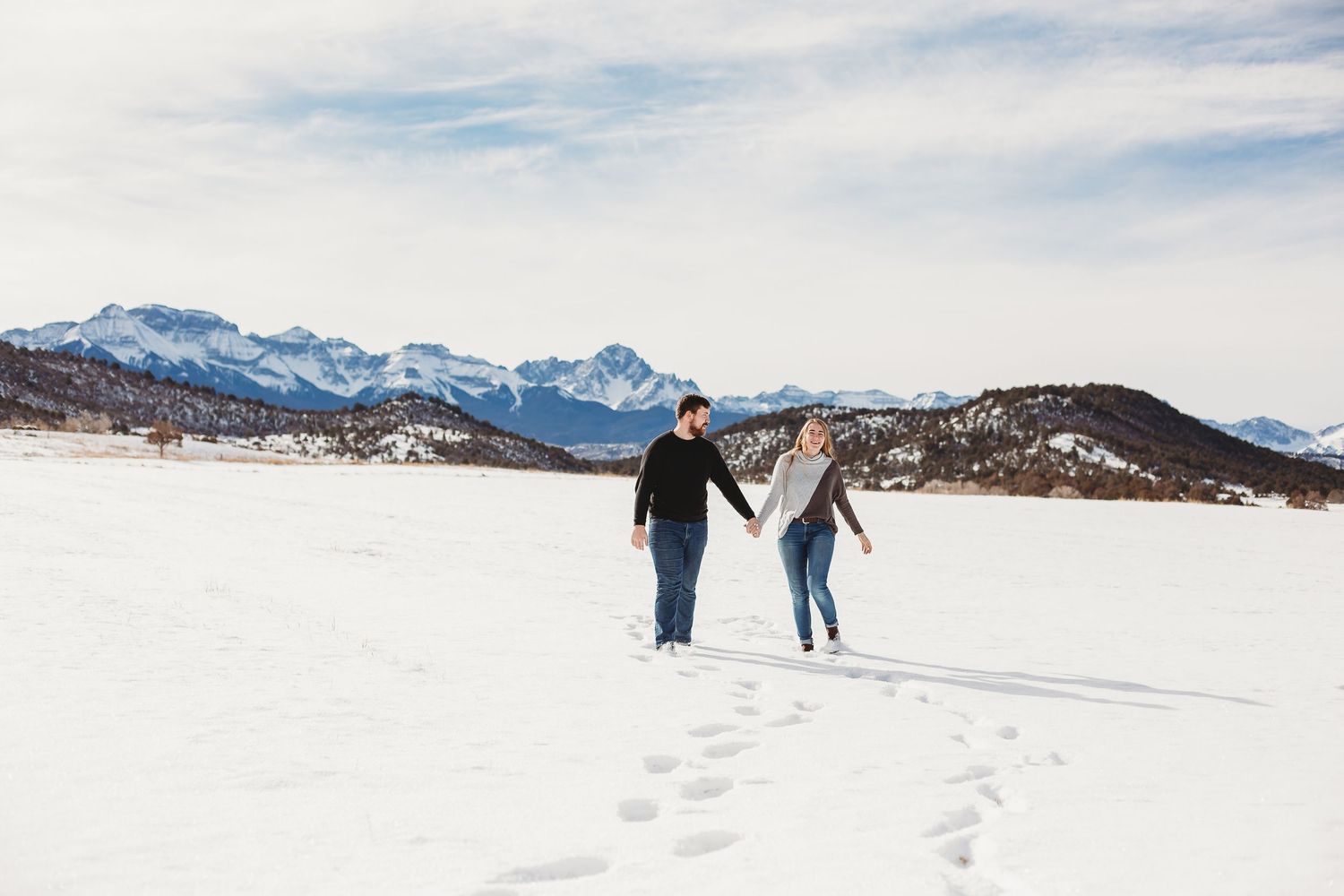 A couple walks together leaving footprints in the snow with mountains on the horizon.
