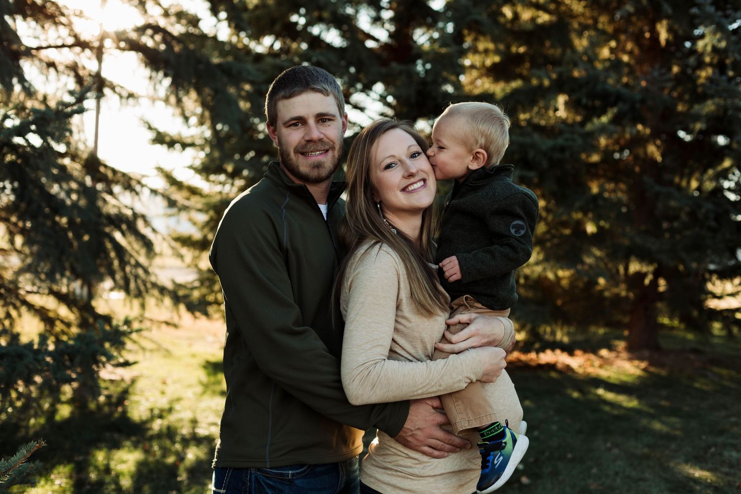 Happy family laughs together during maternity photo session with evergreen trees in background.