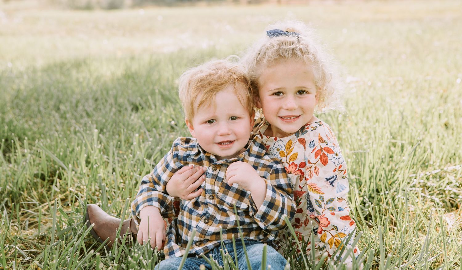 Two children pose together in casual clothes during outdoor photo session in grass.