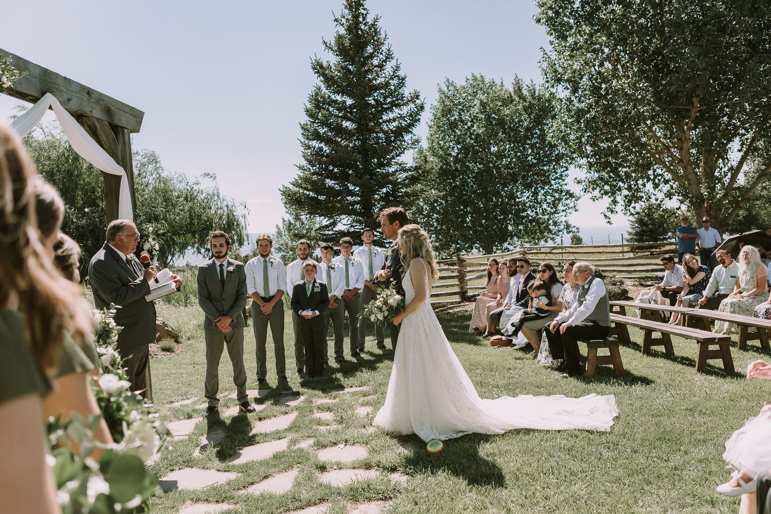 Garden wedding ceremony with guests seated on wooden benches under blue sky.