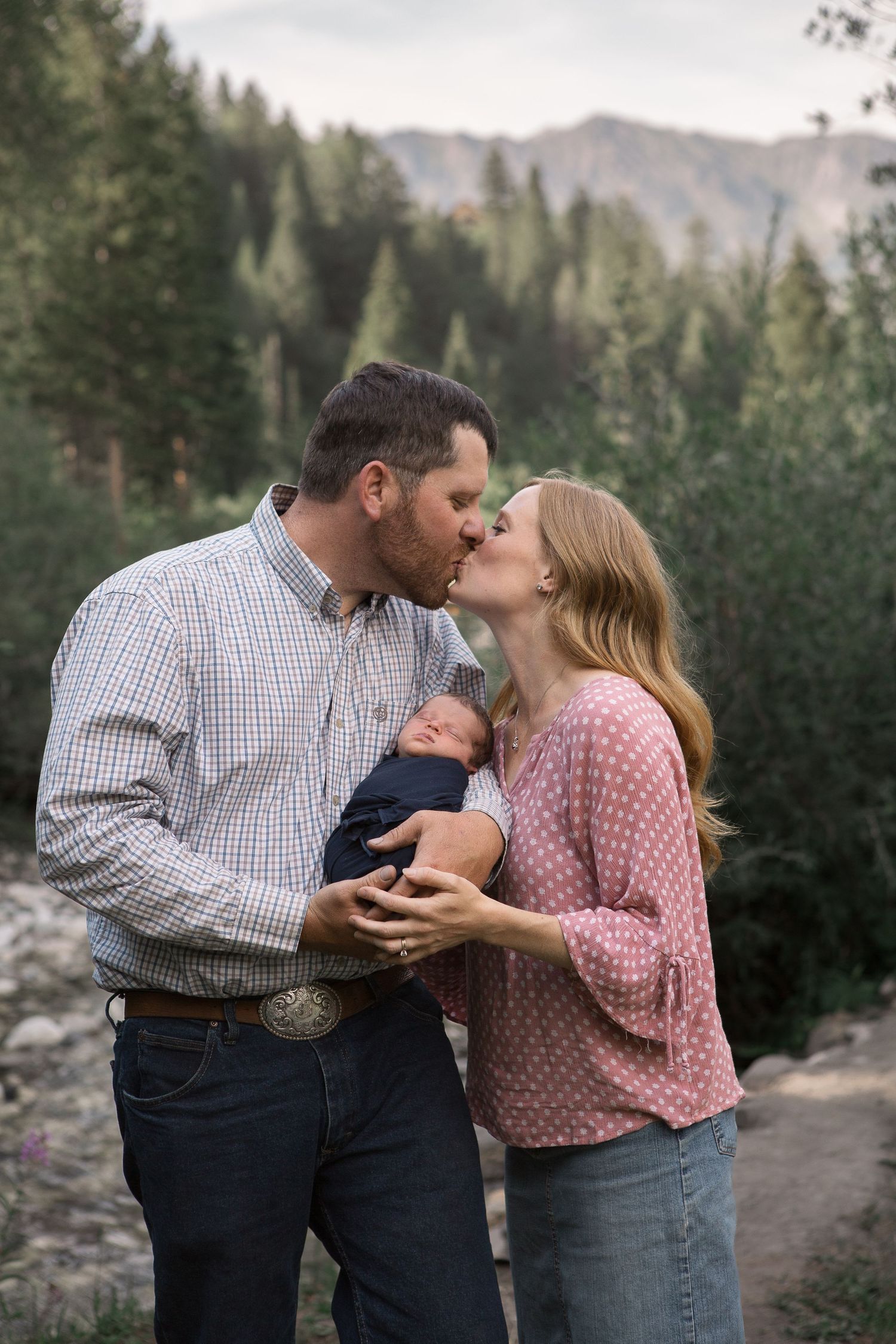 A couple shares an intimate moment with their newborn baby on a mountain trail at sunset.