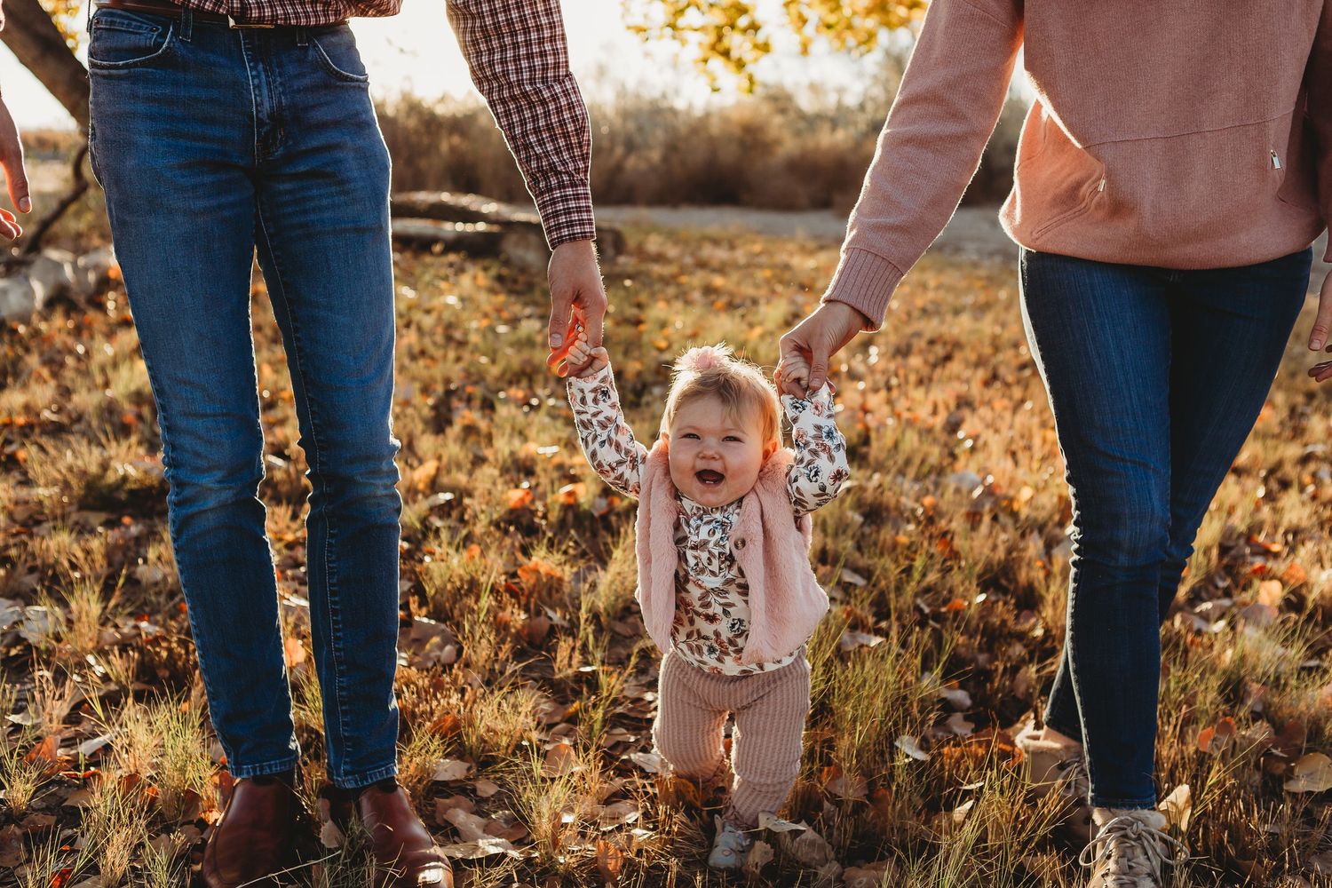 A toddler in pink clothing holds hands with adults while walking through golden autumn leaves at sunset.