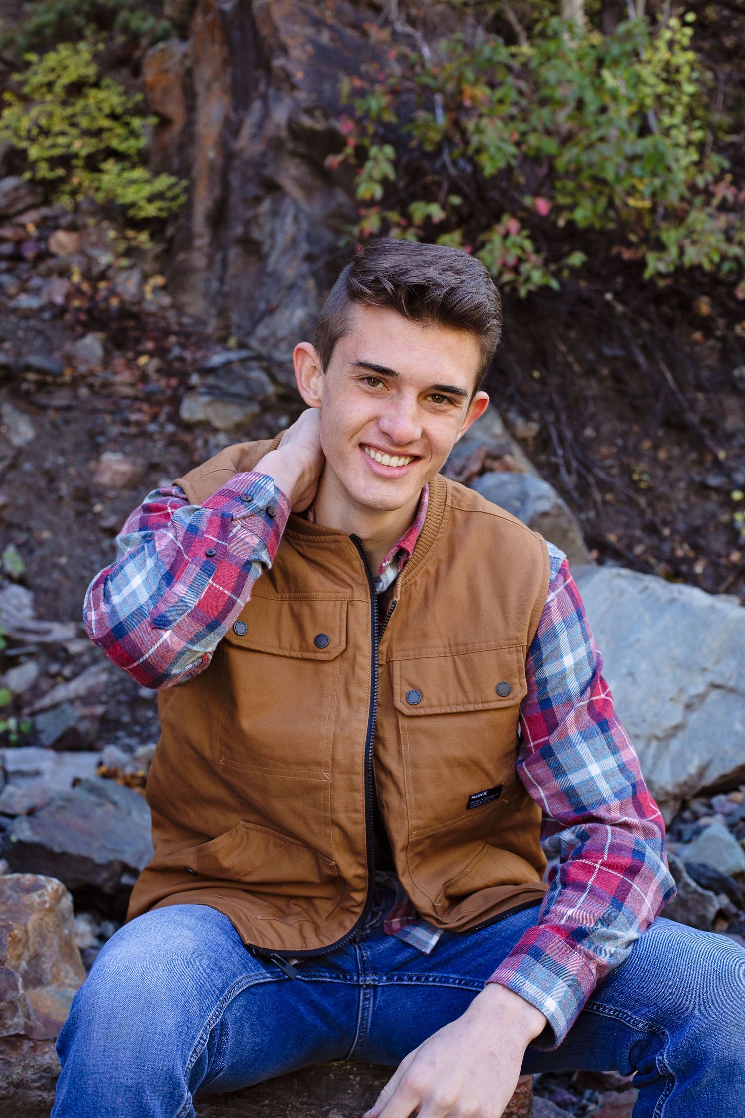 A person in plaid and brown vest sits on rocky terrain with mountains in background.