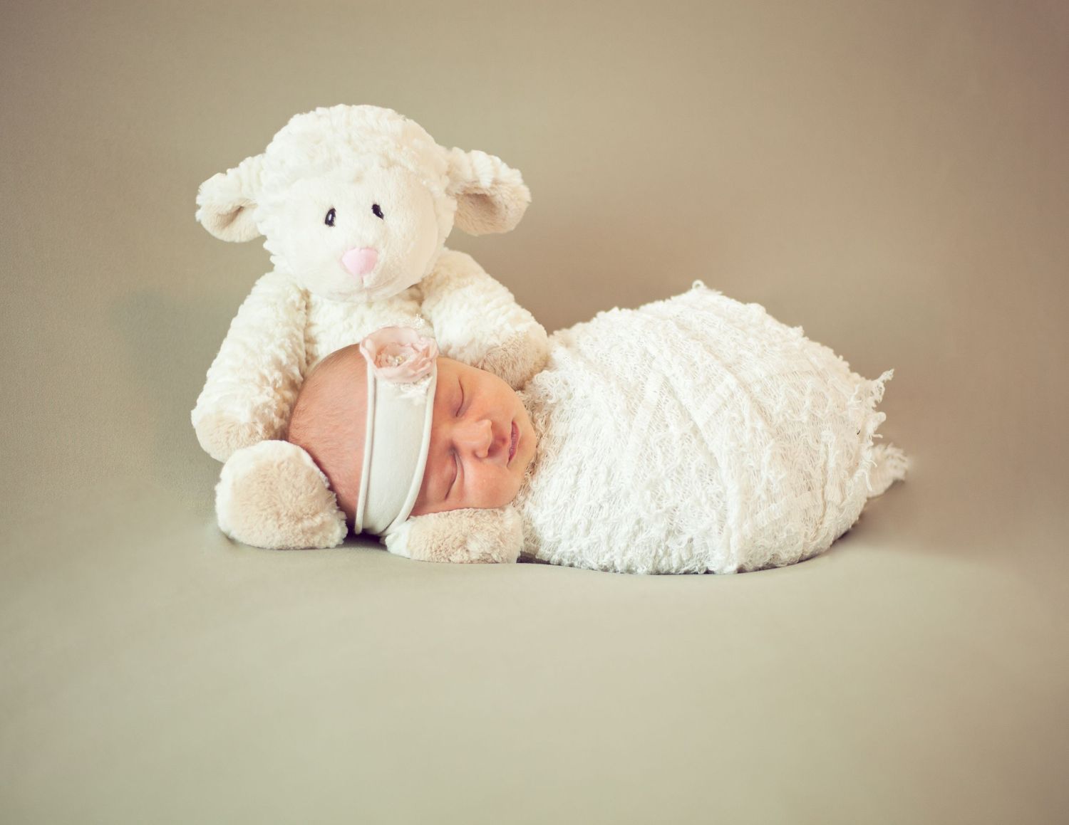 Sleeping newborn posed with plush lamb toy against neutral beige background.