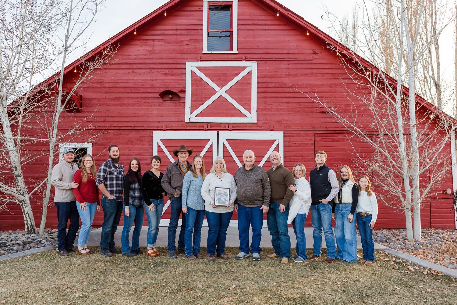 A large group stands in front of a bright red barn with white trim and X-shaped doors on a winter day.