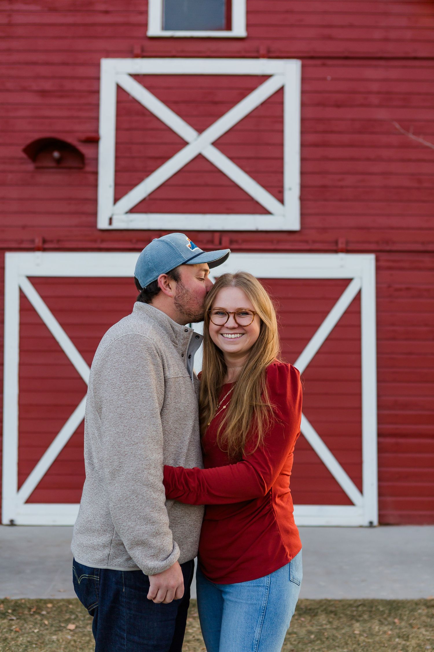 Affectionate couple sharing a moment against backdrop of red barn with white X-pattern doors.