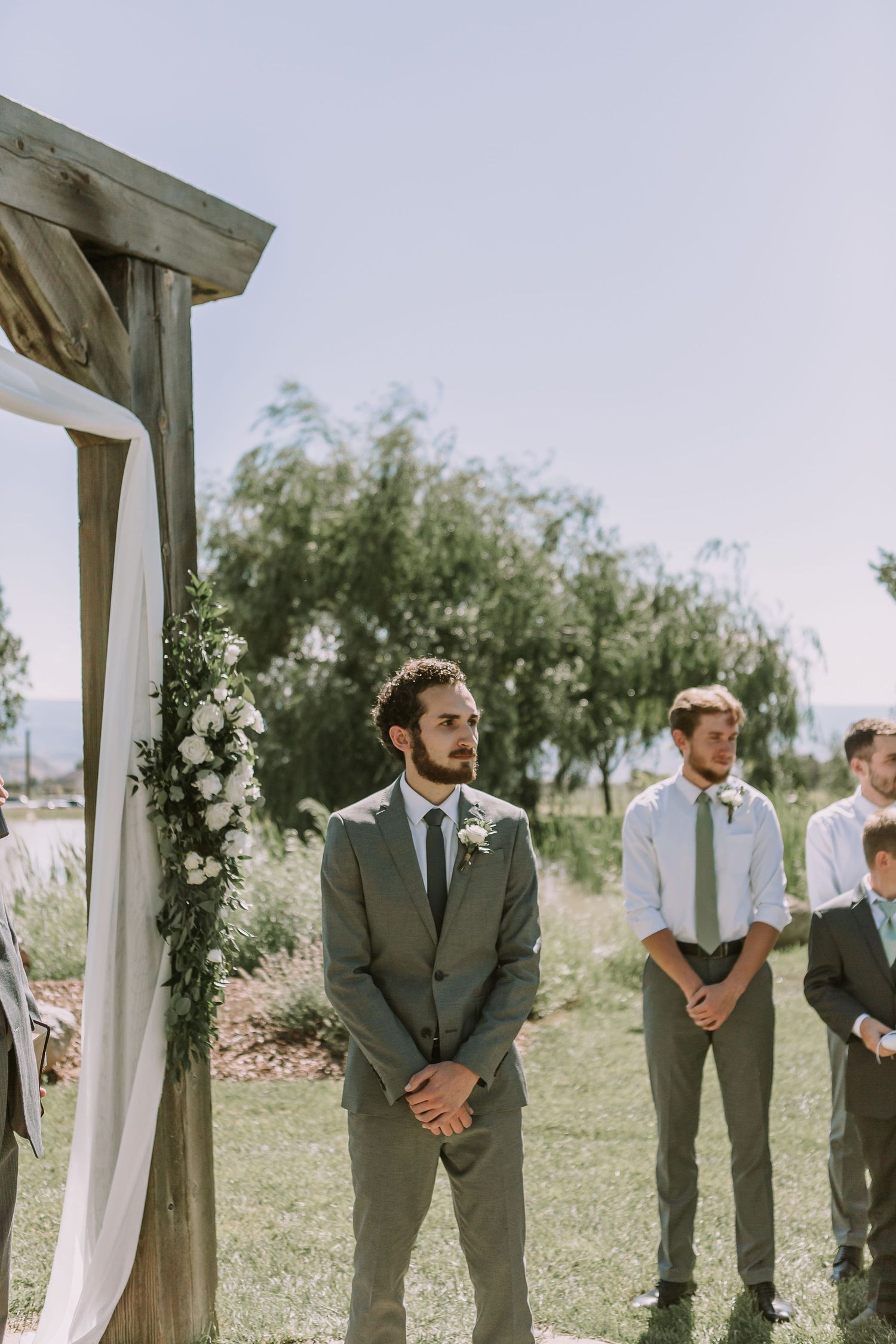 Groom and groomsmen standing at decorated outdoor wedding arch.