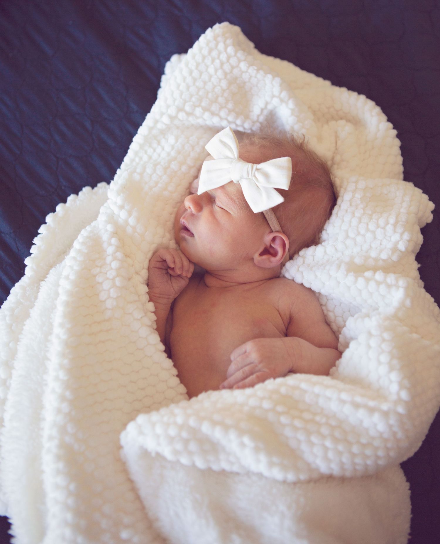 Sleeping newborn wrapped in white textured blanket wearing a white bow headband.