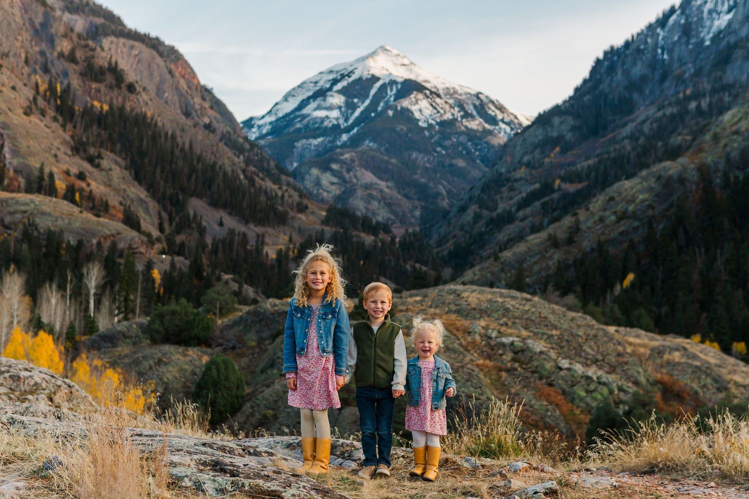 Three siblings pose on a mountain trail with autumn colors and snow-capped peaks in the distance.
