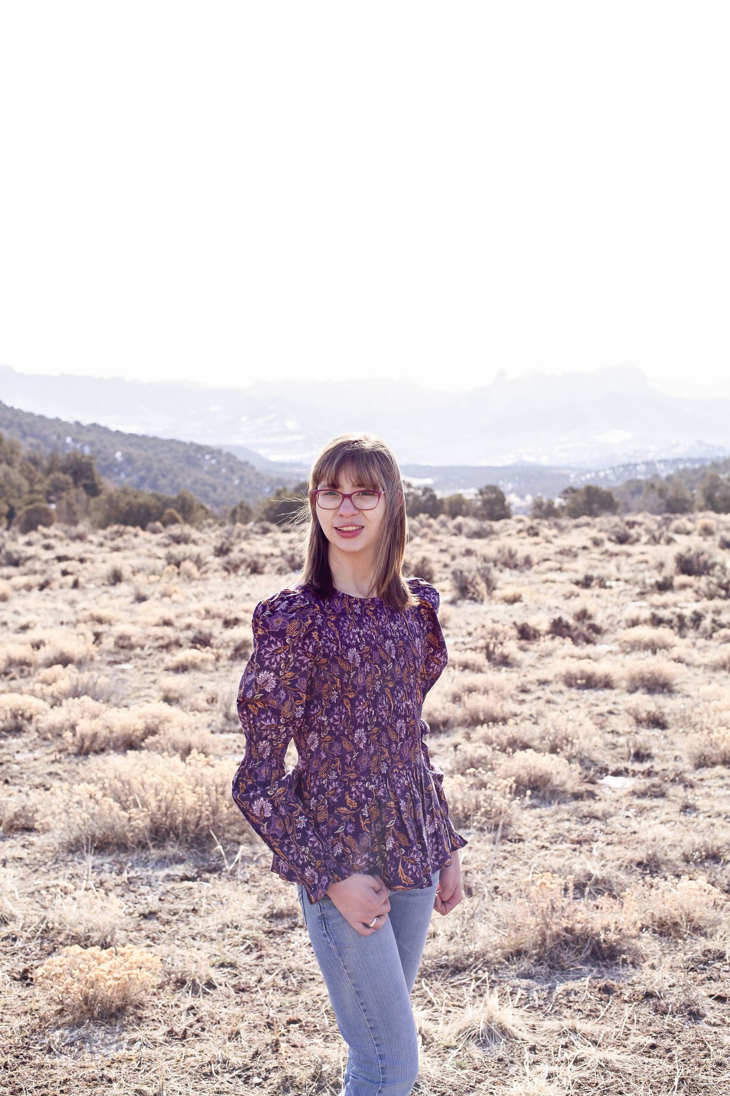 Person in floral top poses in desert landscape with mountains in background.
