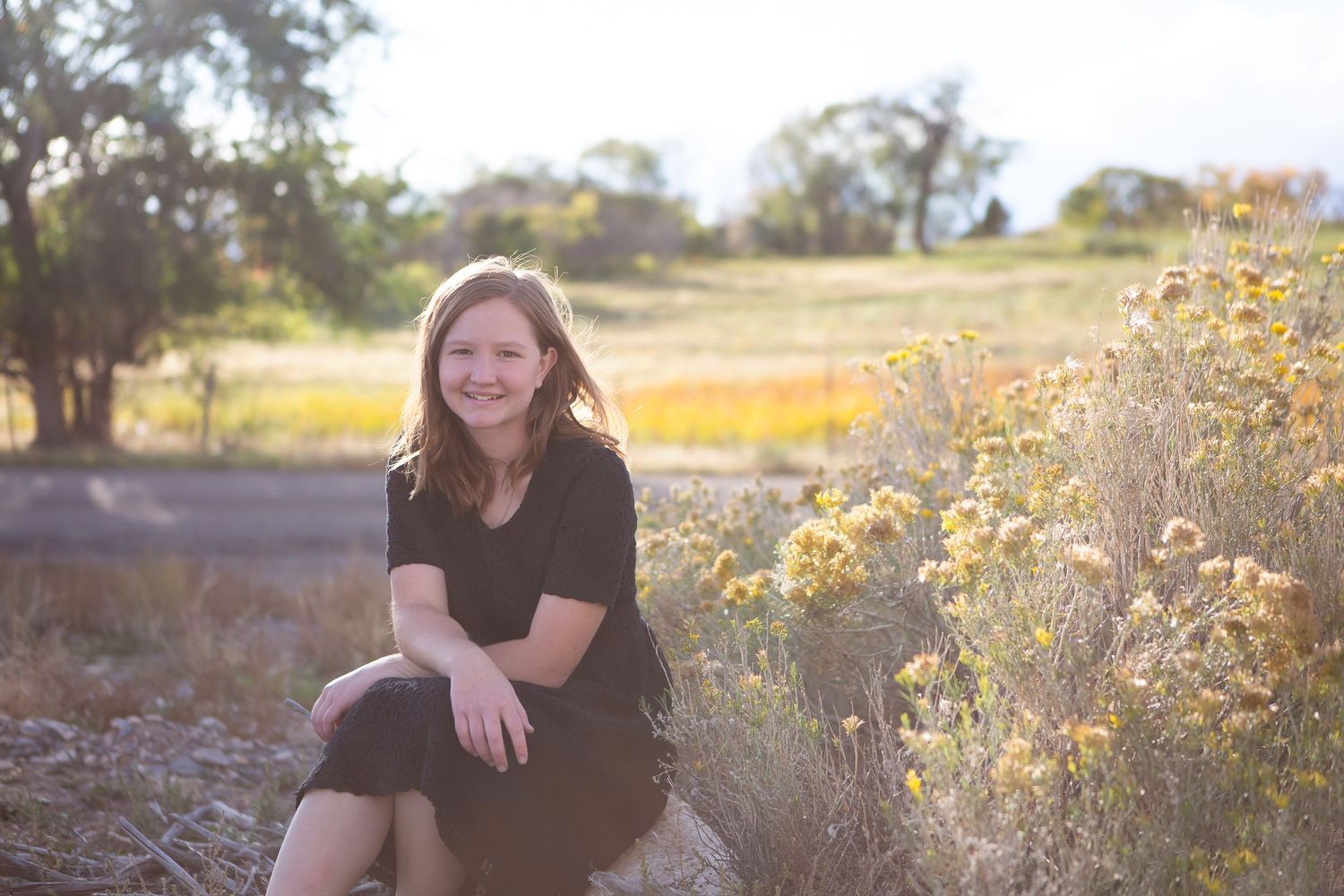 Individual in black dress sits among wildflowers during golden hour in rural setting.