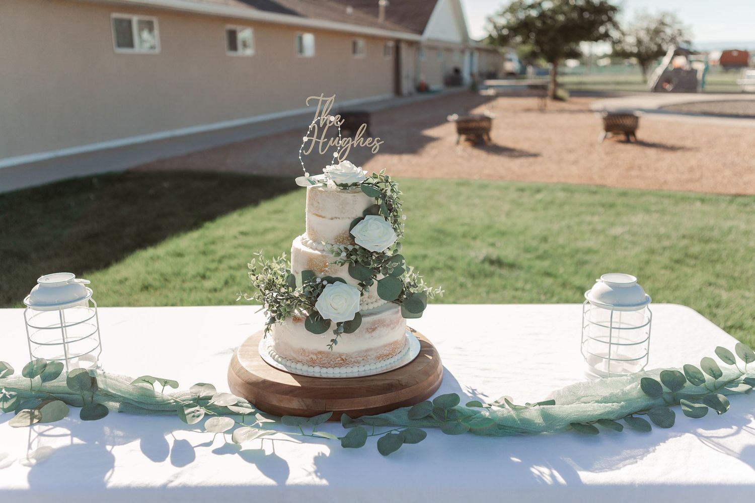 White wedding cake with eucalyptus decor and mason jar lanterns on an outdoor table.