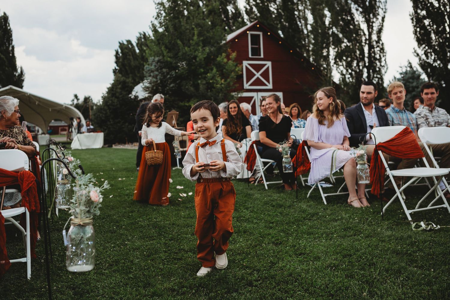 Young attendant walks down grassy aisle at outdoor wedding with red barn in background.