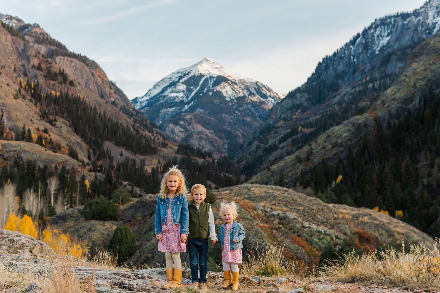 Three children stand together on a mountain trail with a snow-capped peak and fall colors in the valley.
