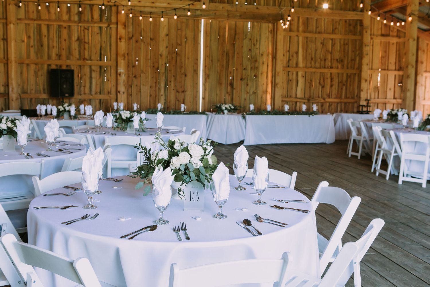 White round tables with folding chairs set for wedding reception under string lights in barn venue.