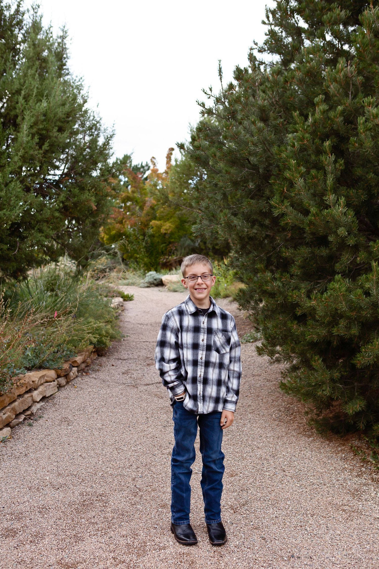 Standing figure on gravel path surrounded by evergreen trees in casual attire.