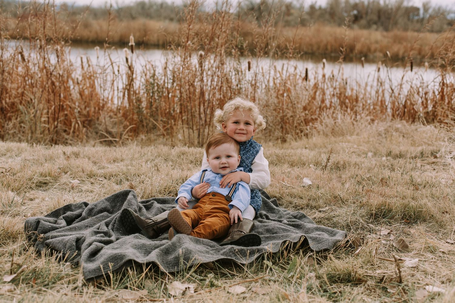 Two children sit on a gray blanket near a pond surrounded by autumn grasses.