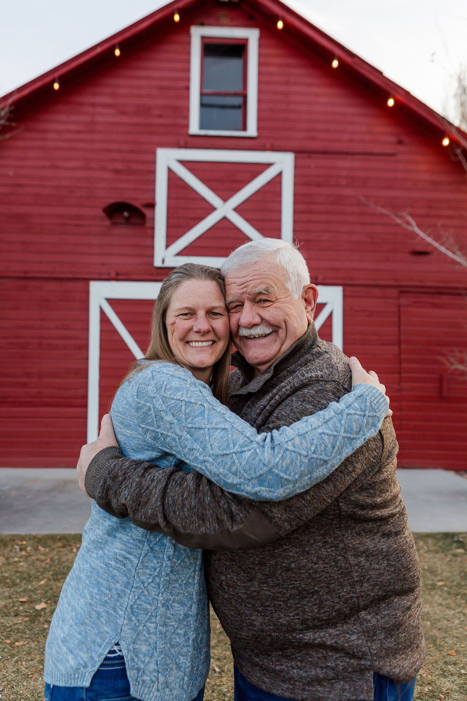 Two people sharing a joyful hug in front of a bright red barn with white decorative trim.