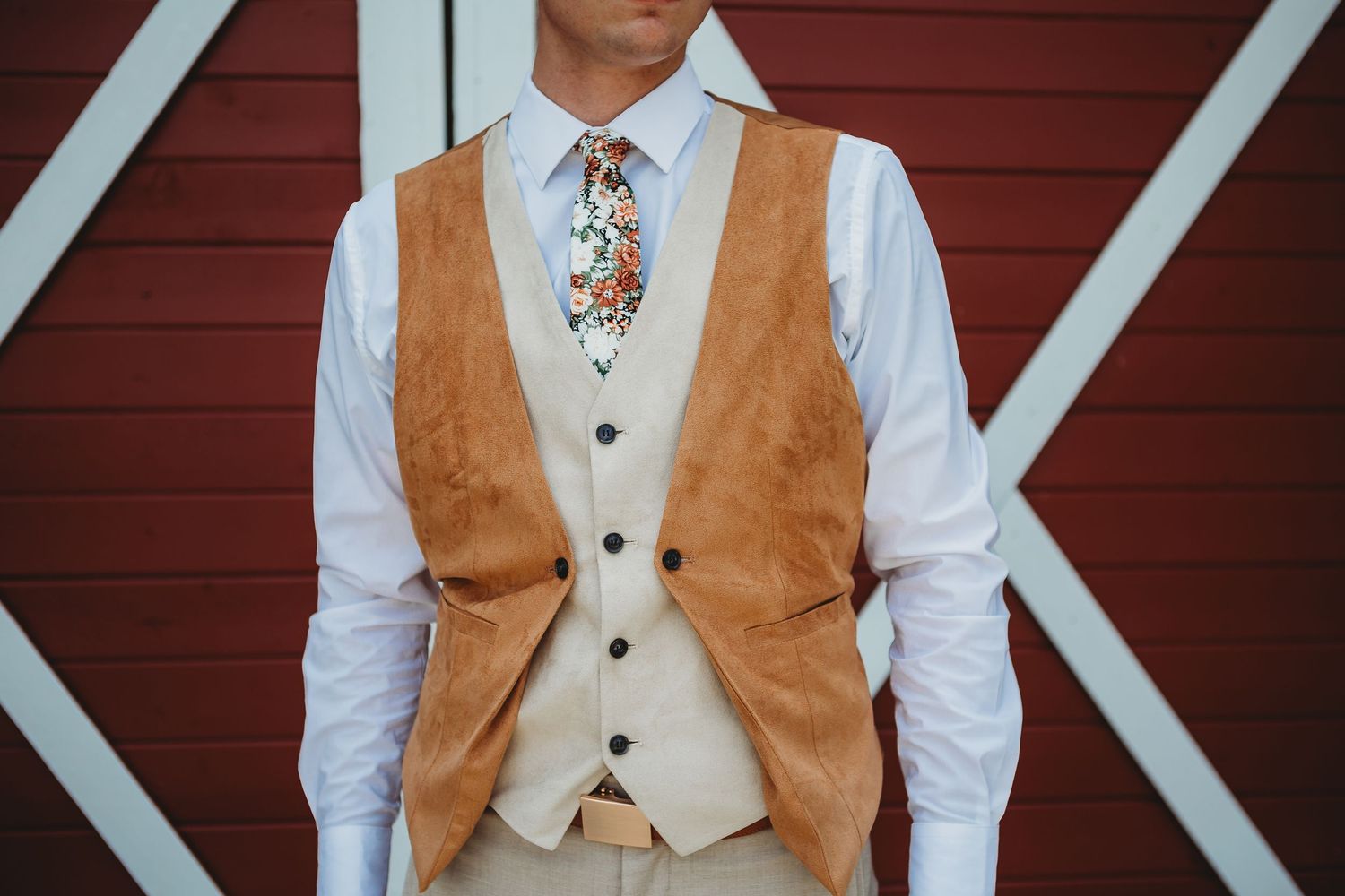 Groom's attire featuring a tan suede vest, white shirt, and floral tie against red barn doors.