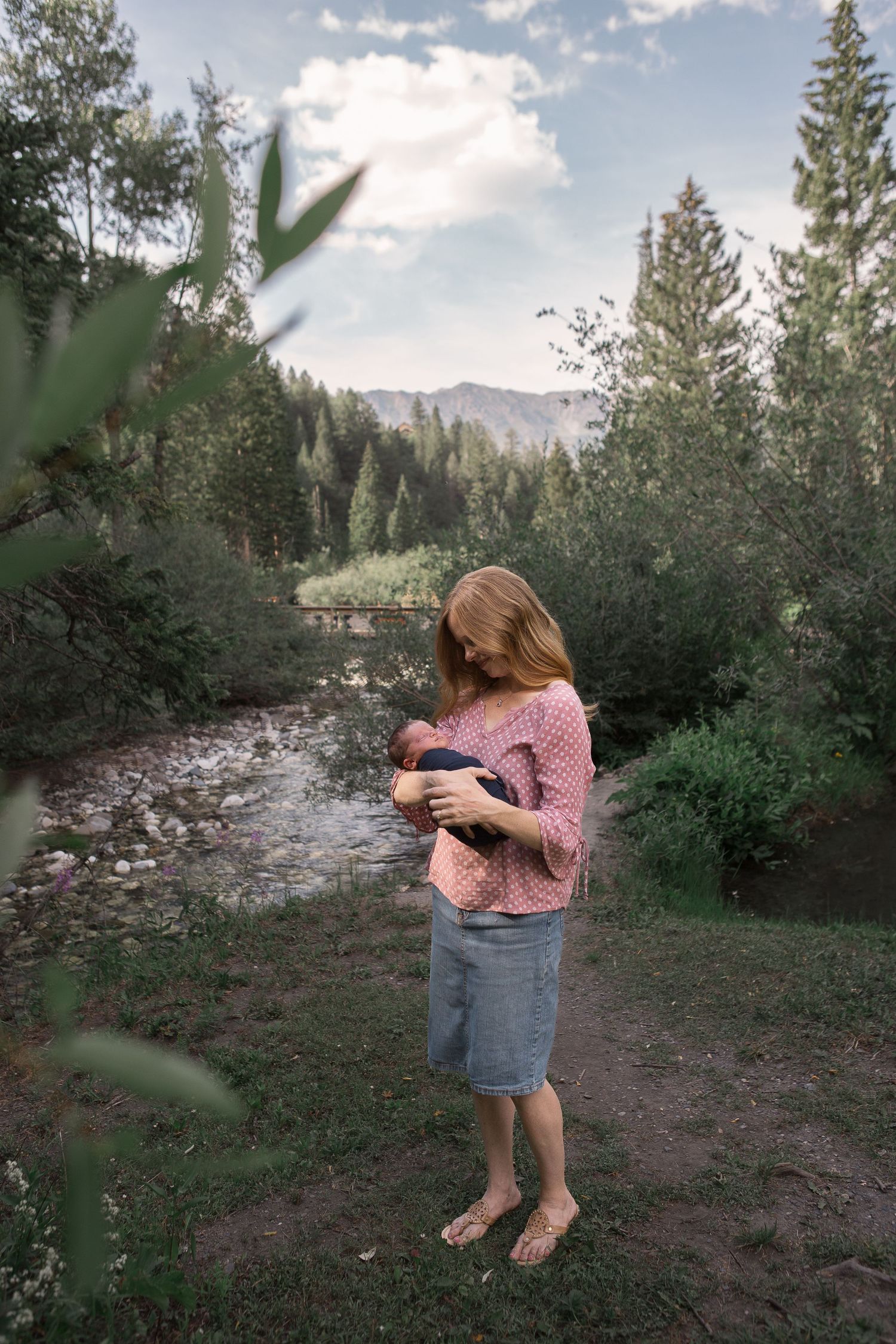 A person holds a baby while standing barefoot by a mountain stream surrounded by evergreen trees in nature.