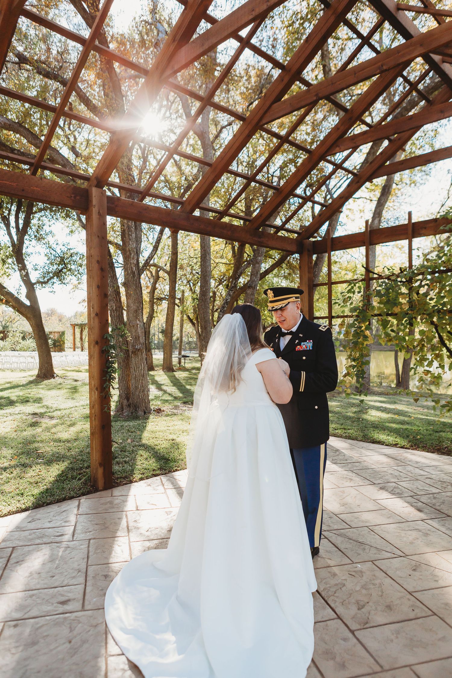 Wedding couple embraces under wooden pergola with sunlight streaming through.