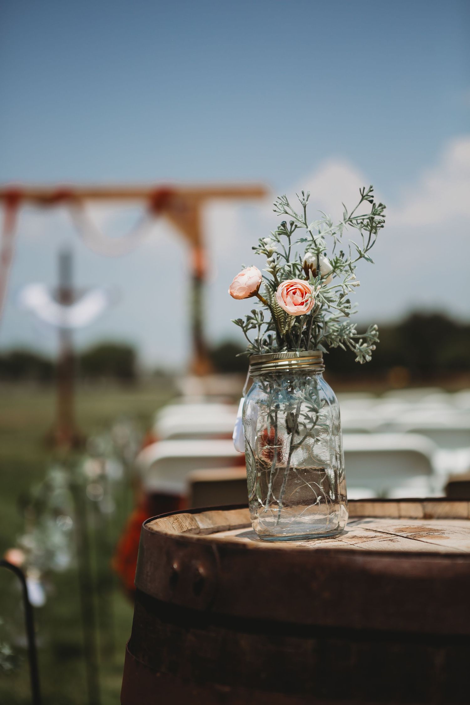 Mason jar with delicate pink roses and baby's breath sits atop rustic wooden barrel at outdoor wedding venue.