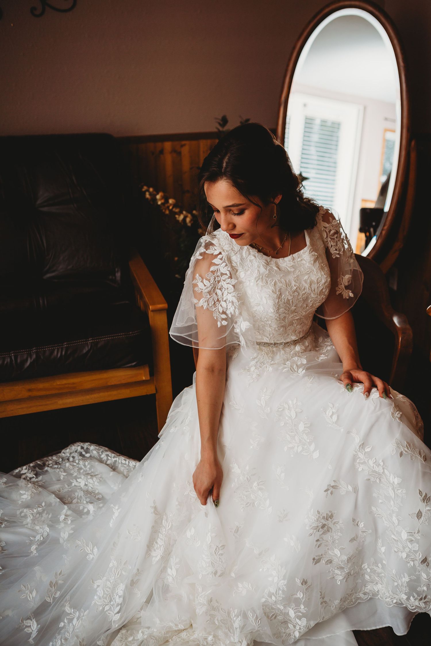 Bride in lace sleeve wedding gown sits gracefully next to vintage mirror.