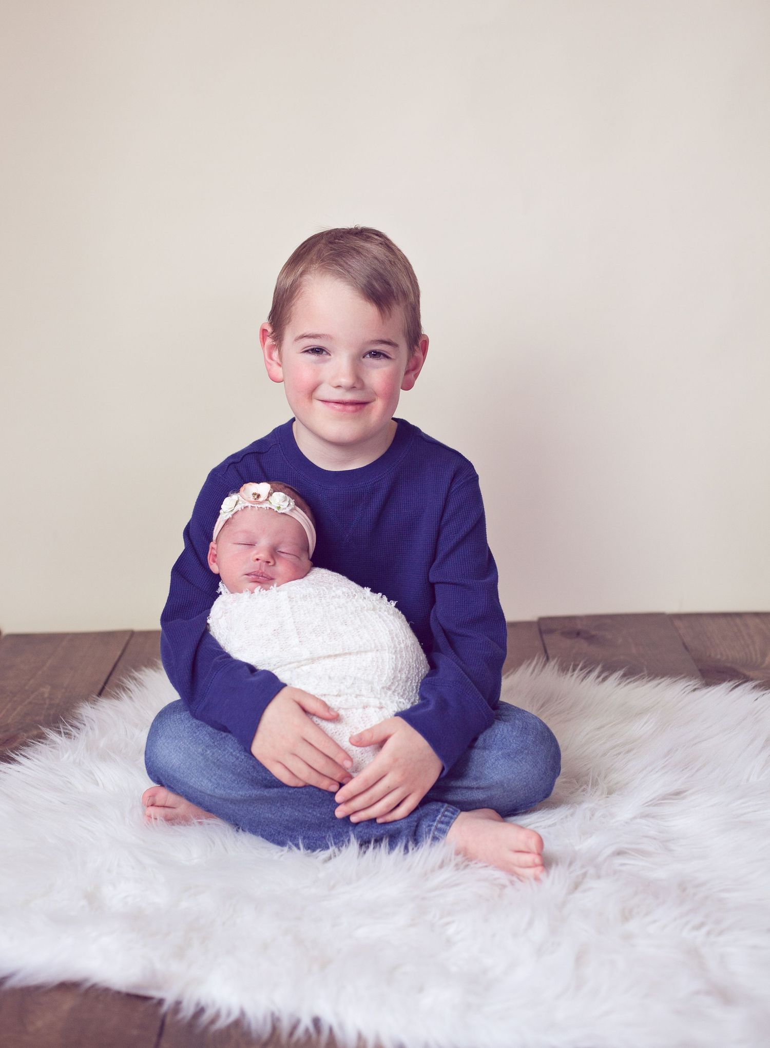 A sibling portrait on a white furry rug with a swaddled newborn.