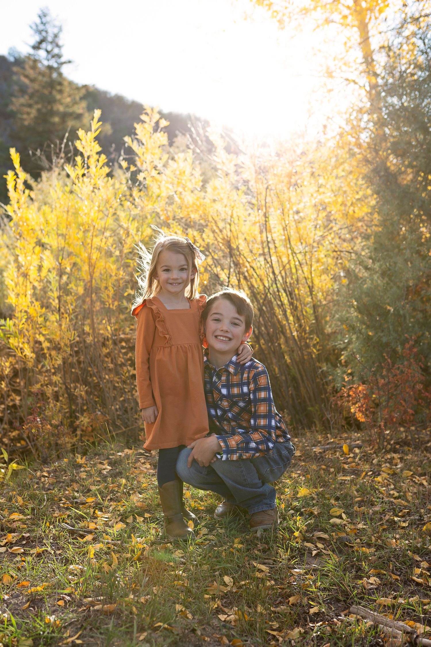 Two children pose together in autumn foliage with golden sunlight streaming through.
