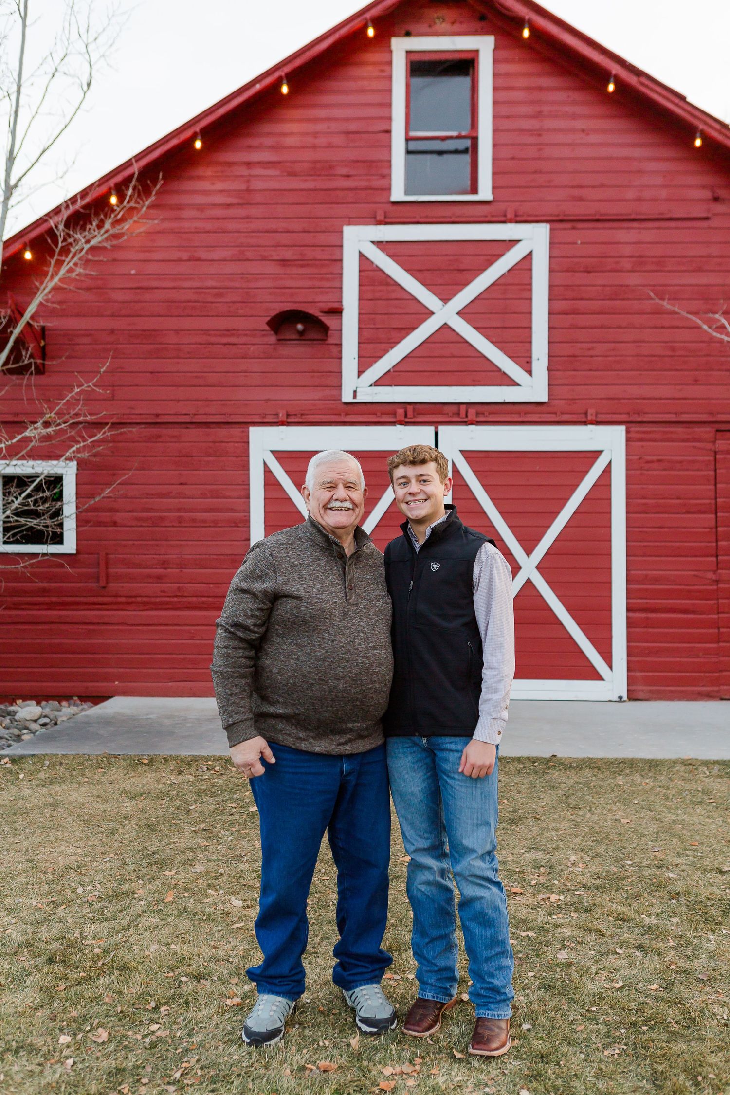 Two people stand side by side in front of a classic red barn with white X-pattern trim.