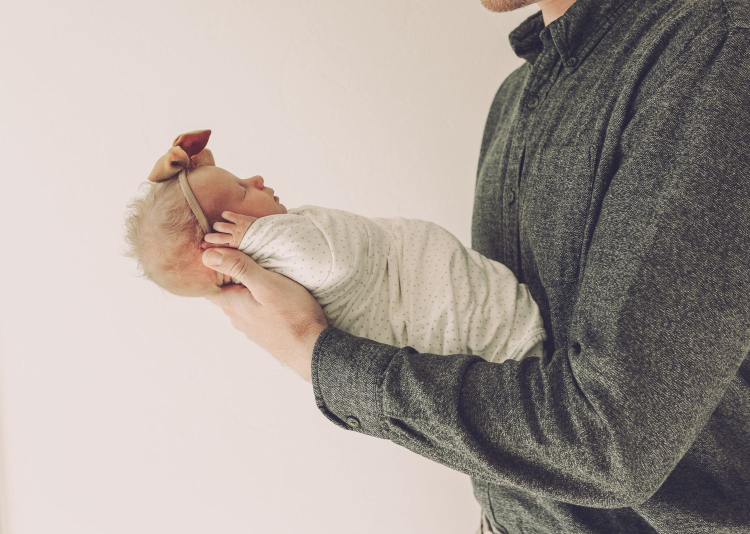 Side view of person in grey sweater holding swaddled infant against neutral background.