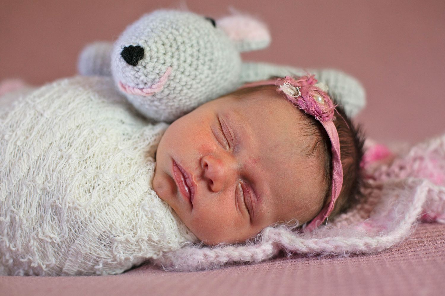 Sleeping newborn with pink headband next to knitted stuffed animal on pink background.
