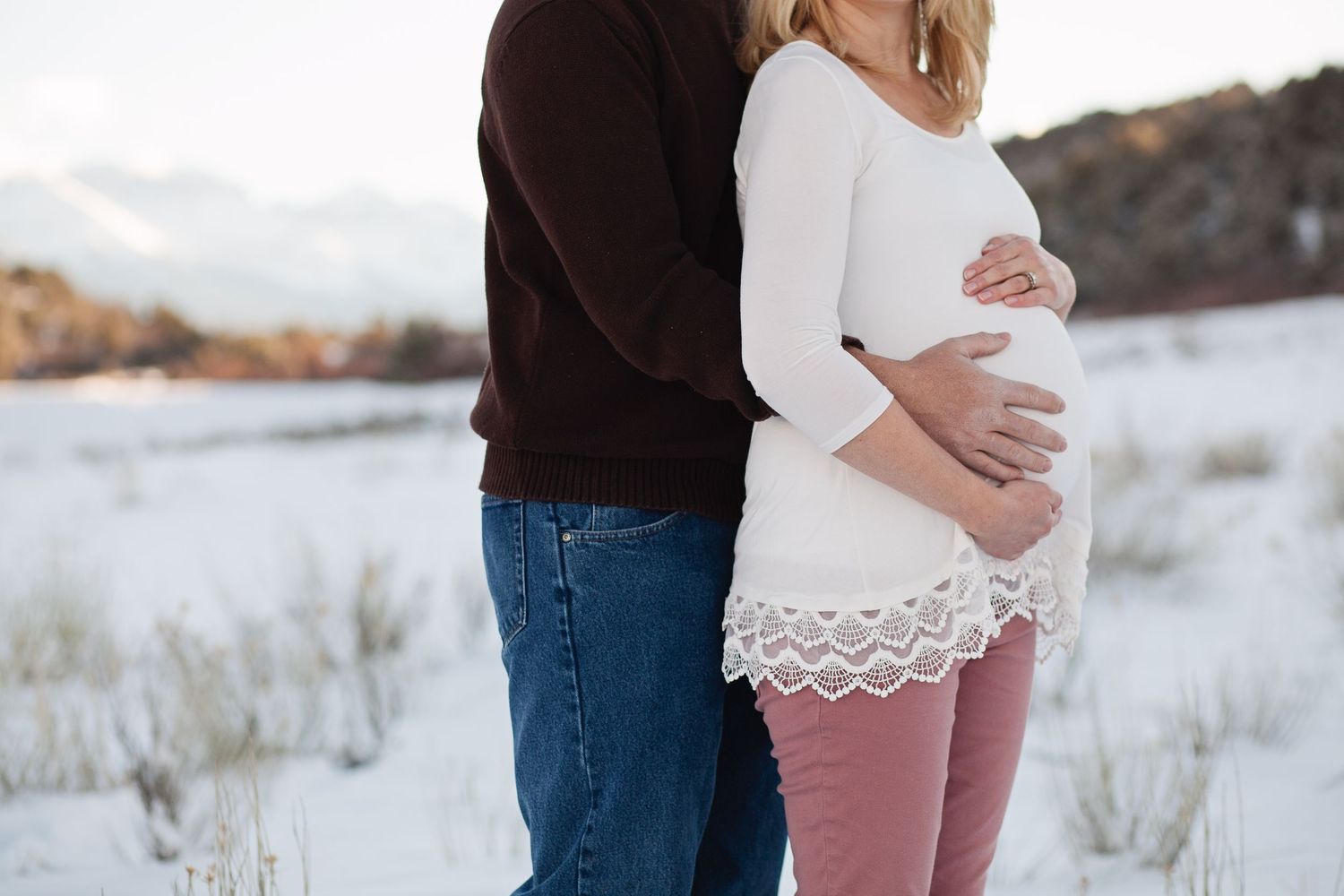 Intimate winter maternity photo with couple in snow against mountain backdrop.