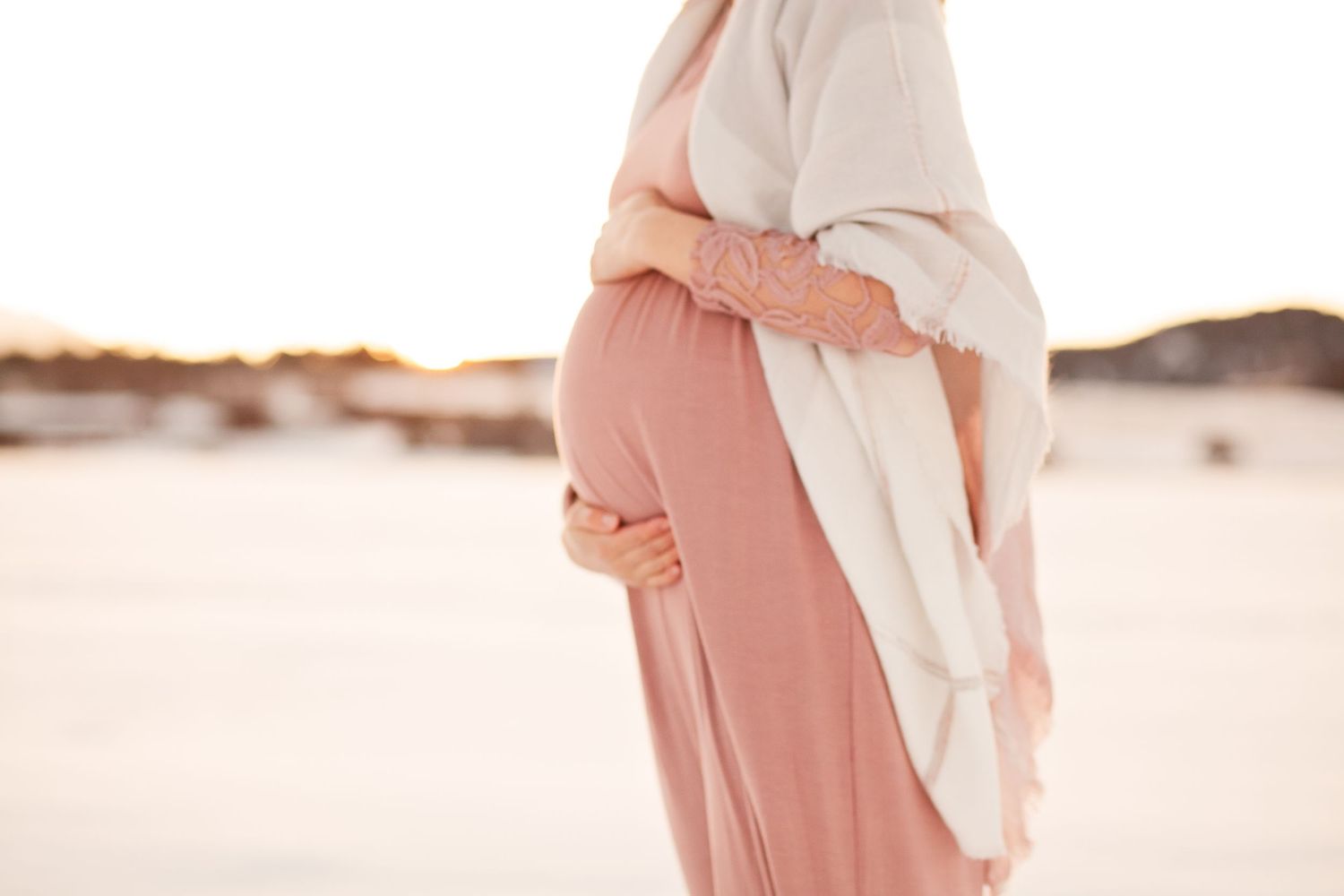 Artistic cropped photo of pregnant woman in flowing pink dress against snowy background.