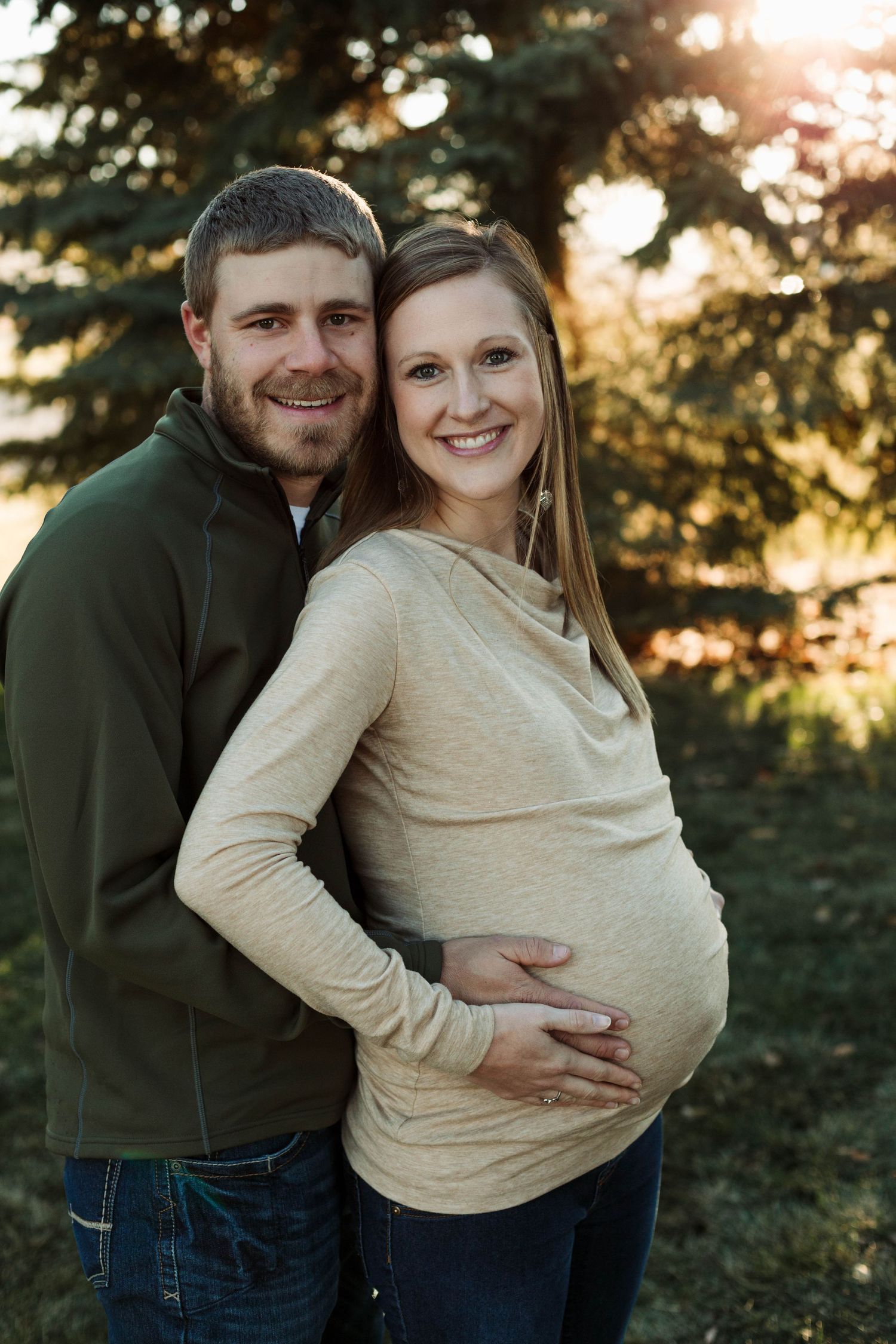 Loving couple poses for maternity photo in front of evergreen trees at golden hour.