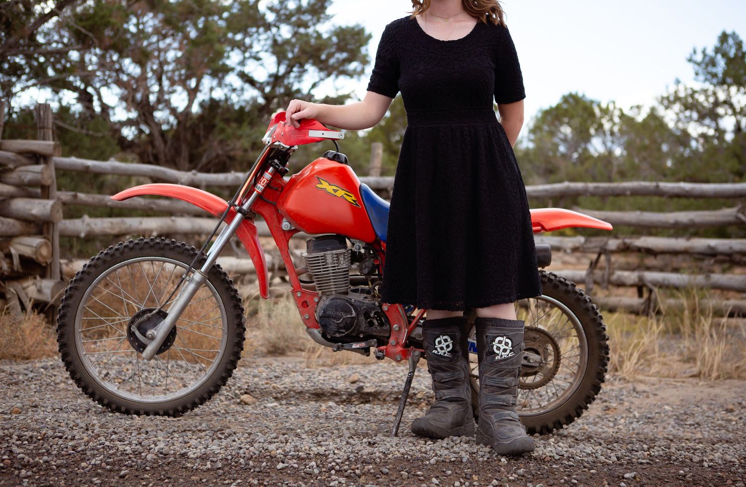A person in a black dress stands next to a vintage red dirt bike on a dusty trail.