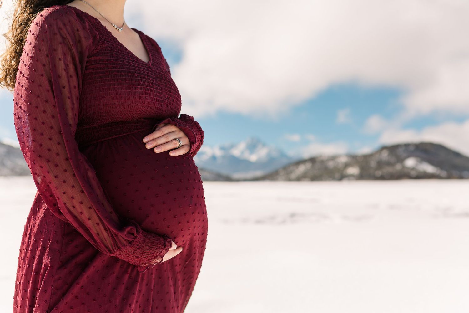 A maternity photo showing a burgundy dress against a snowy mountain landscape.