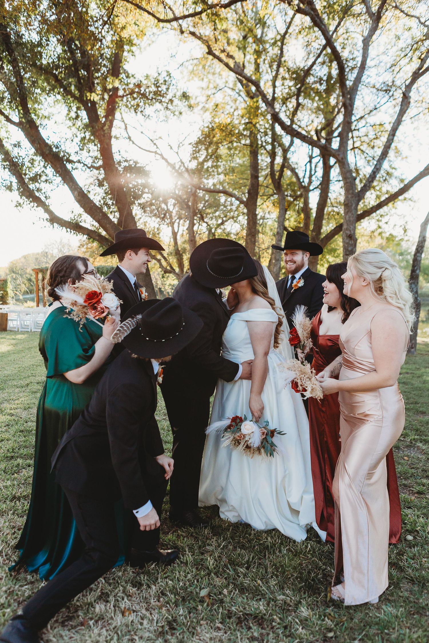 A wedding party gathers under trees during golden hour for a celebratory group photo.