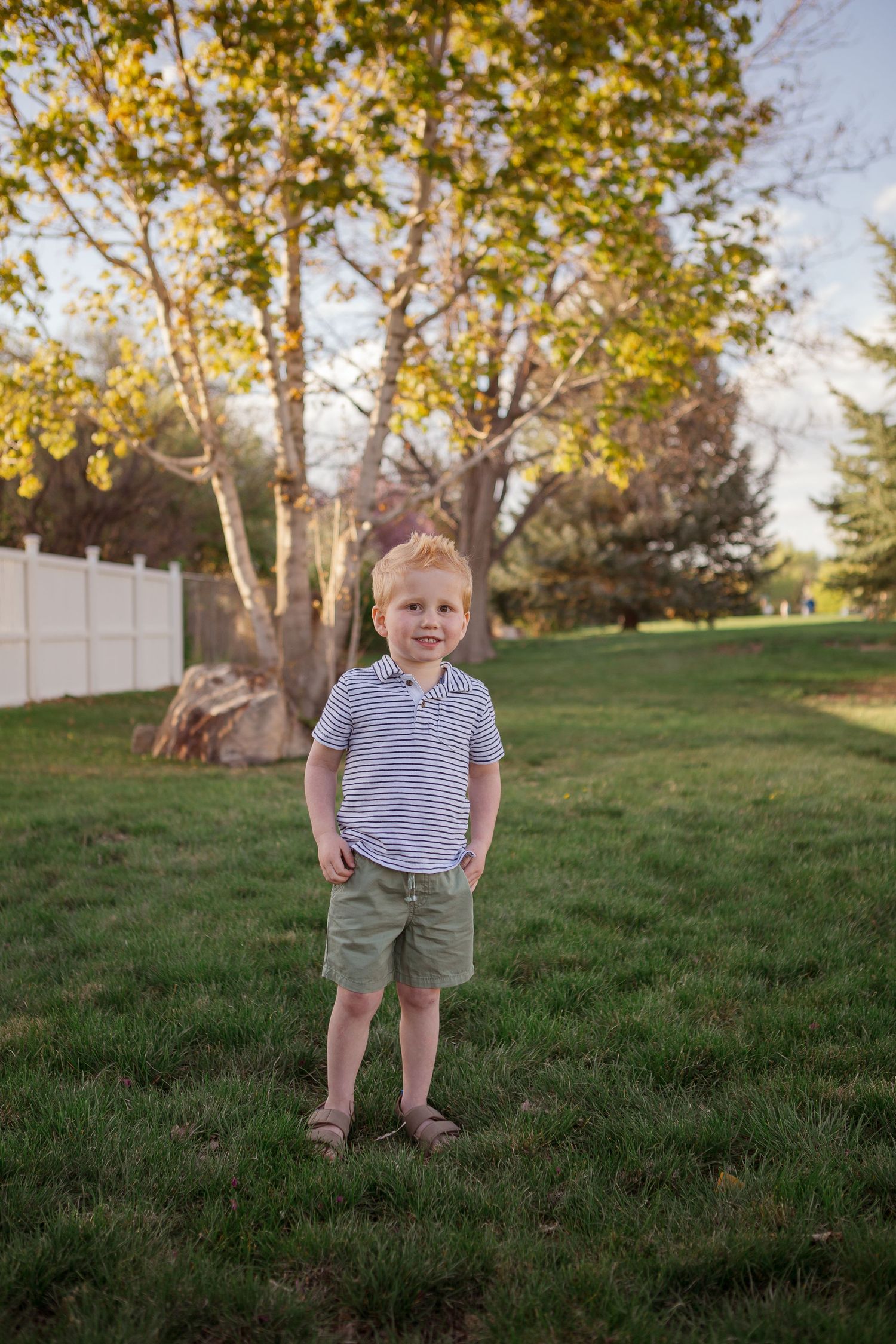 A young child in striped shirt and shorts standing on grass with trees in the background.