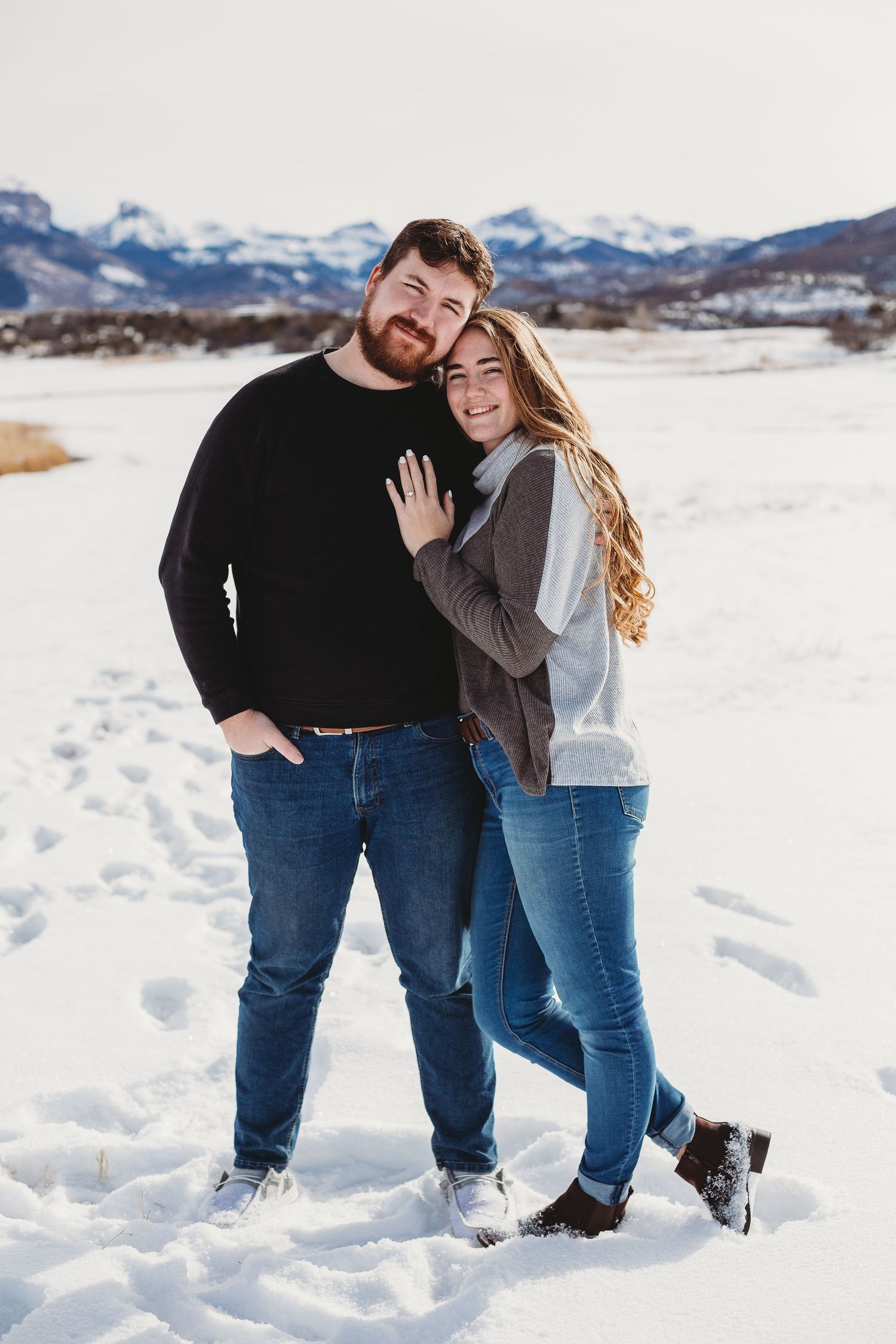 A couple poses together in casual winter clothing against a snowy mountain landscape.