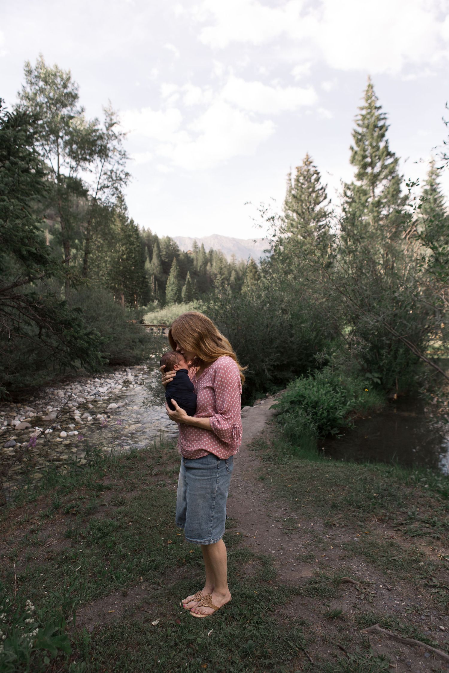 Pink-clad figure holding infant on a scenic mountain trail with rushing creek and evergreen trees.