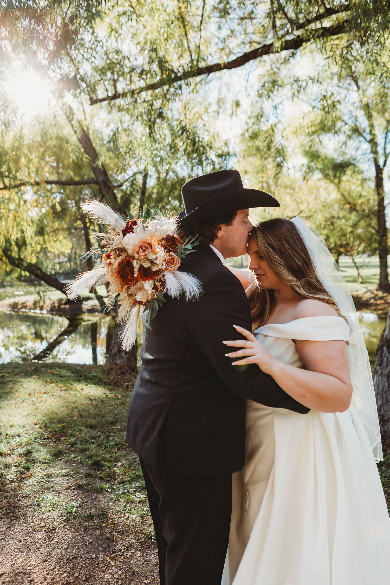 Couple in wedding attire share intimate moment under sunlit trees with boho bouquet.
